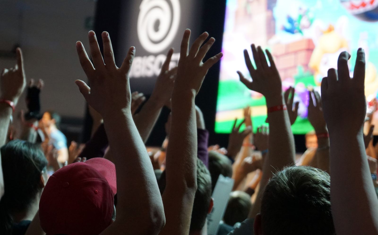 A crowd of fans raising their hands toward a big screen at a busy game booth at gamescom