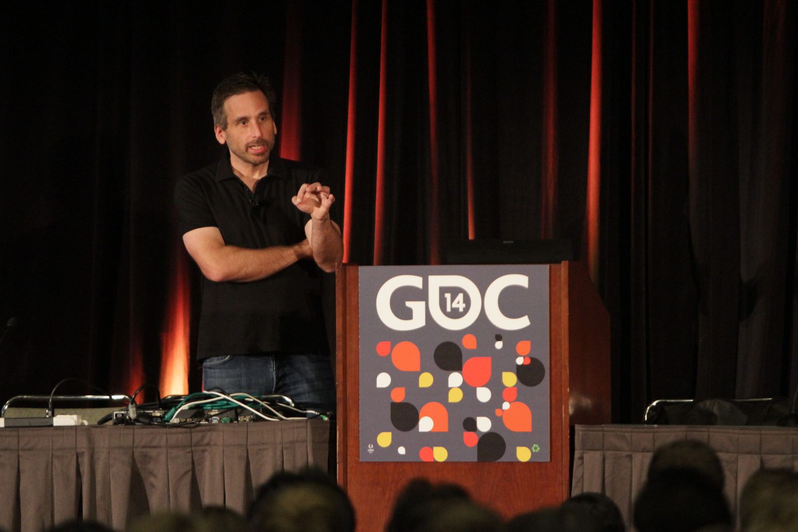 BioShock creative director Ken Levine speaking beside a GDC 14 podium in front of a dark red curtain