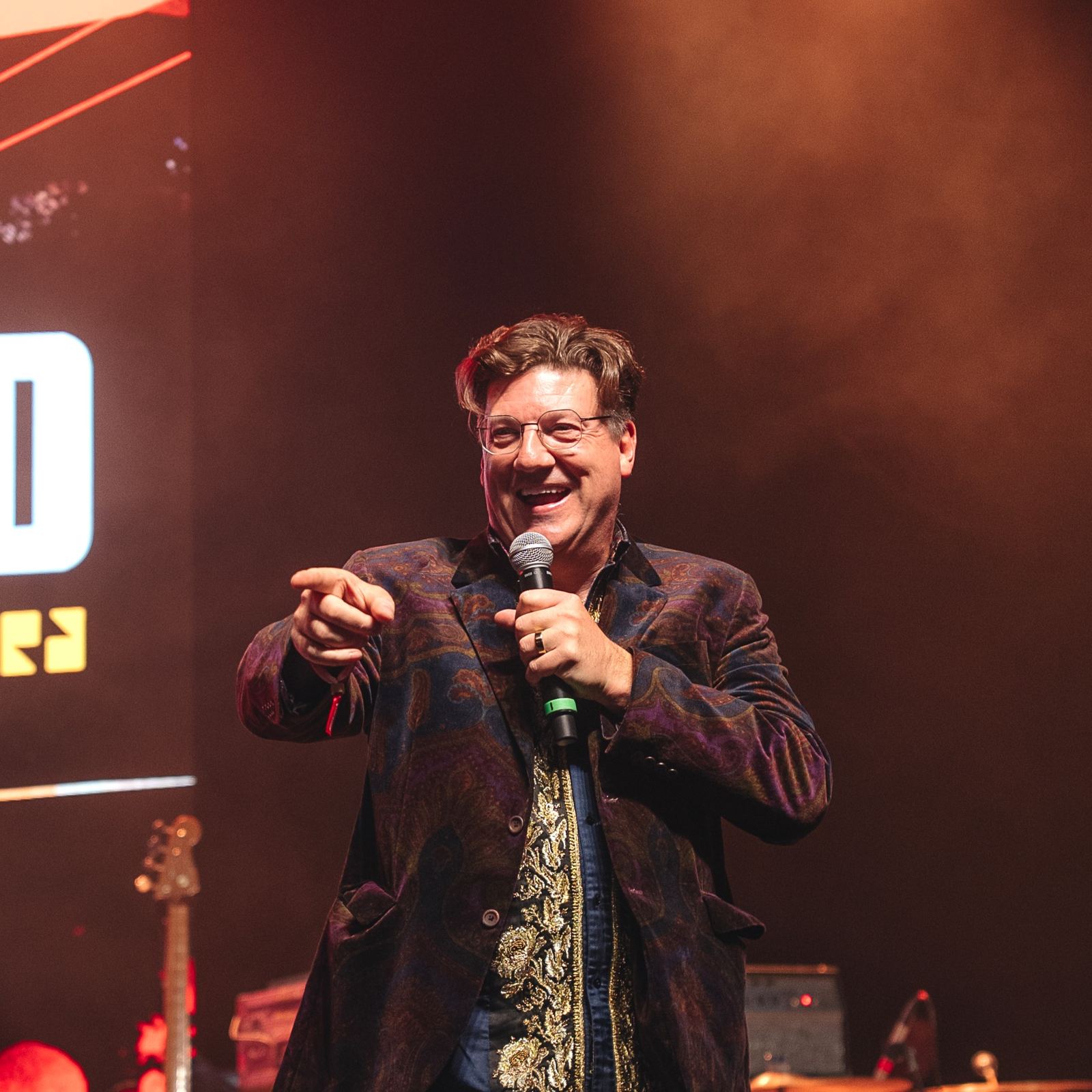 Gearbox founder Randy Pitchford on stage with a microphone, smiling and pointing at the crowd under red stage lights