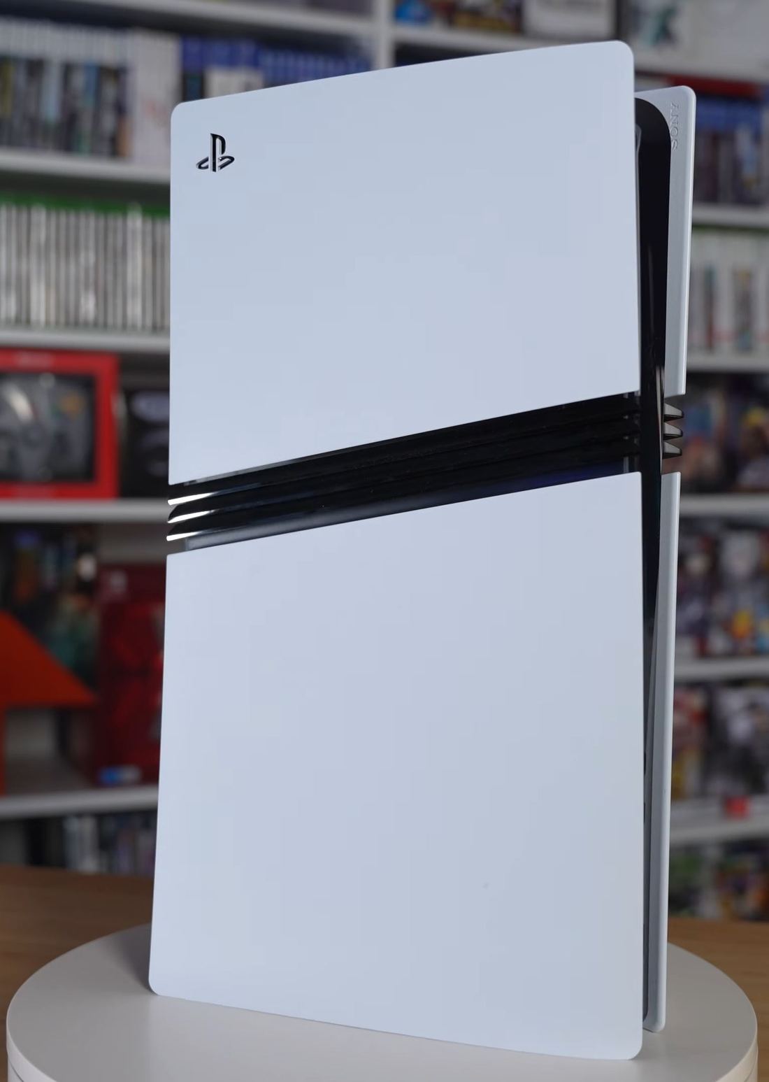 A white PlayStation 5 Pro standing upright on a desk in front of shelves of games