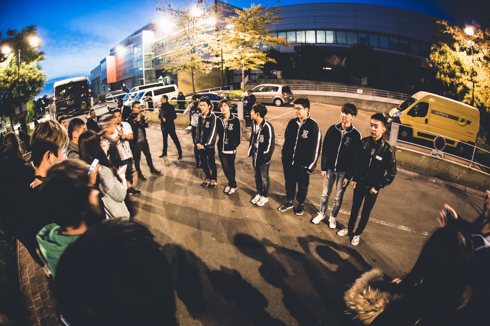 A team of esports players in matching track jackets lined up outside an arena in Paris at dusk, watched by fans